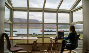 A woman sitting with a laptop, overlooking a tranquil lake surrounded by majestic mountains. She appears focused on the screen, symbolizing virtual mental health care accessibility in serene, natural settings.