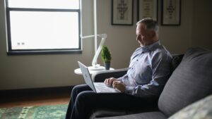 A man sitting comfortably on a couch at home, engaged in a virtual psychological appointment. He is focused on his laptop screen, showcasing the convenience of virtual mental health consultations